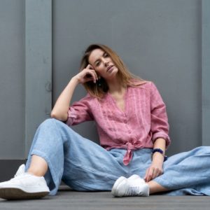 Kako nositi najmodernije bluze ovog leta; žena sedi na podu u širokim plavim pantalonama, belim patikama i modernoj roze bluzi vezanoj u čvor